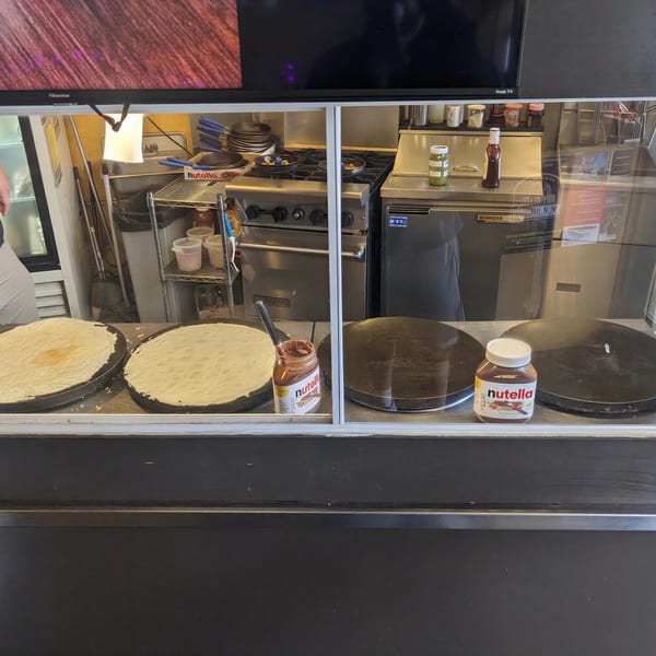 Crepes cooking on griddles with jars of Nutella and kitchen equipment visible behind a glass counter.