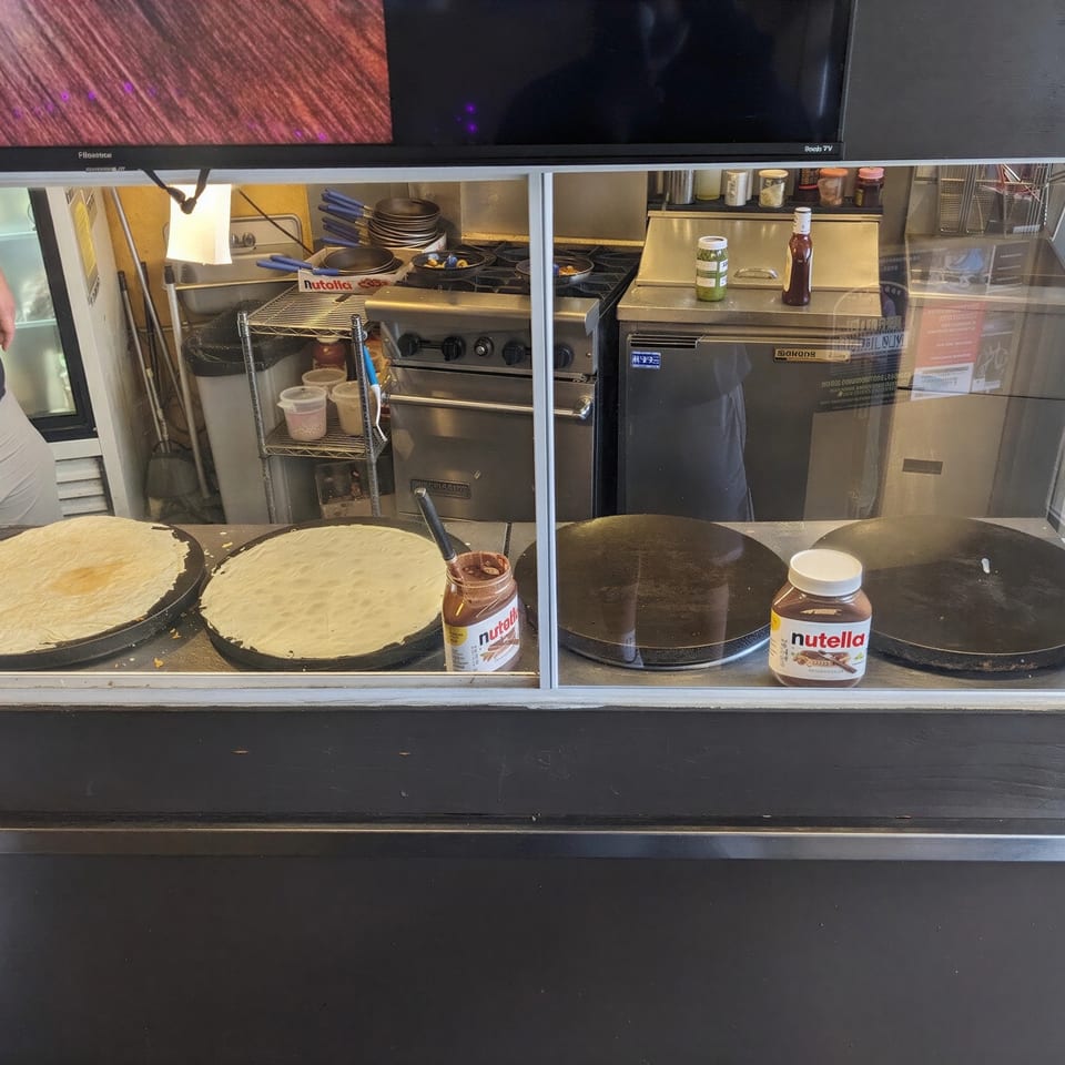 Crepes cooking on griddles with jars of Nutella and kitchen equipment visible behind a glass counter.