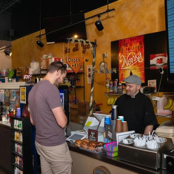 A customer orders from a smiling barista behind a counter with pastries and coffee supplies.