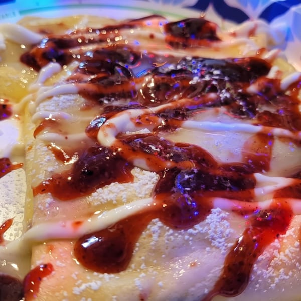 A dessert crepe topped with strawberry sauce, white icing, powdered sugar, and whipped cream.