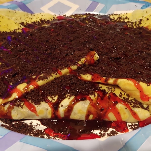 A dessert crepe topped with chocolate crumbles, chocolate syrup, and red fruit drizzle on a paper plate.