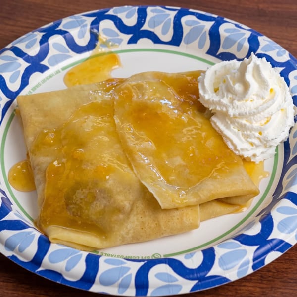 Crepes drizzled with orange sauce and topped with whipped cream on a blue and white paper plate.