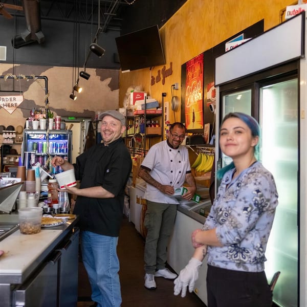 Three smiling restaurant staff members, two men and one woman, stand behind a counter in a busy kitchen.