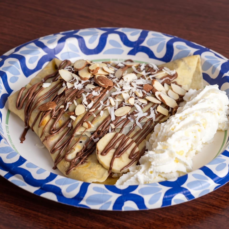Crepe drizzled with chocolate, topped with almonds, coconut flakes, banana slices, and whipped cream.
