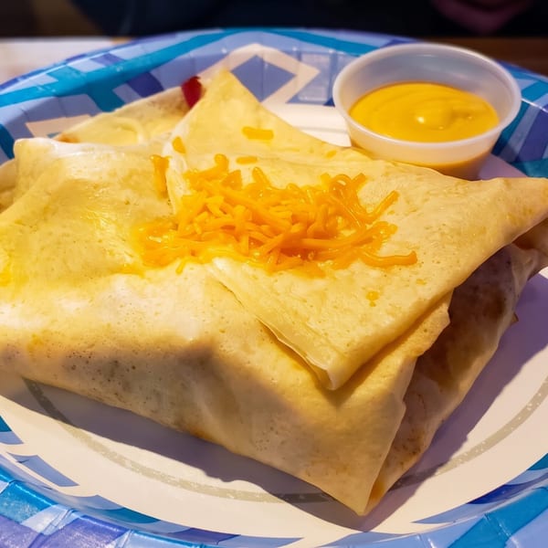 Two burritos topped with shredded cheese and a side of cheese sauce on a blue and white paper plate.