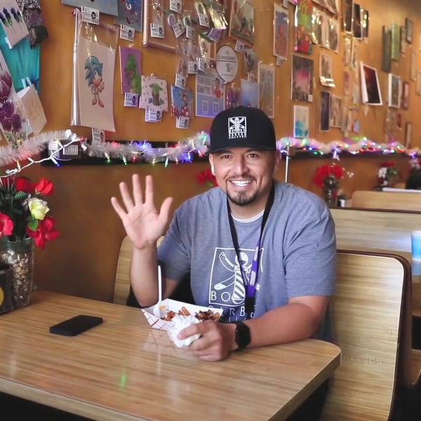 Man in a cap and t-shirt waving, sitting at a booth in a casual restaurant.