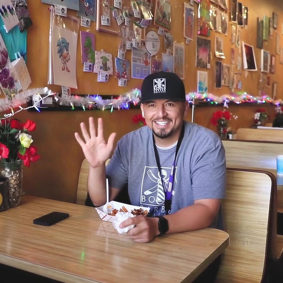 Man in a cap and t-shirt waving, sitting at a booth in a casual restaurant.