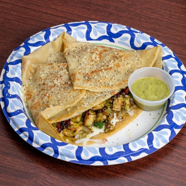 Savory chicken and vegetable crepe with melted cheese and a side of green sauce on a paper plate.