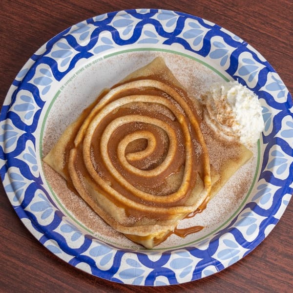 Cinnamon roll crepe with caramel drizzle, cinnamon sugar, and whipped cream on a blue and white plate.